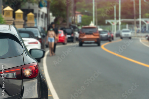 Cars parked in a queue beside the asphalt road. And blurry images of people walking and cars passing by in front.