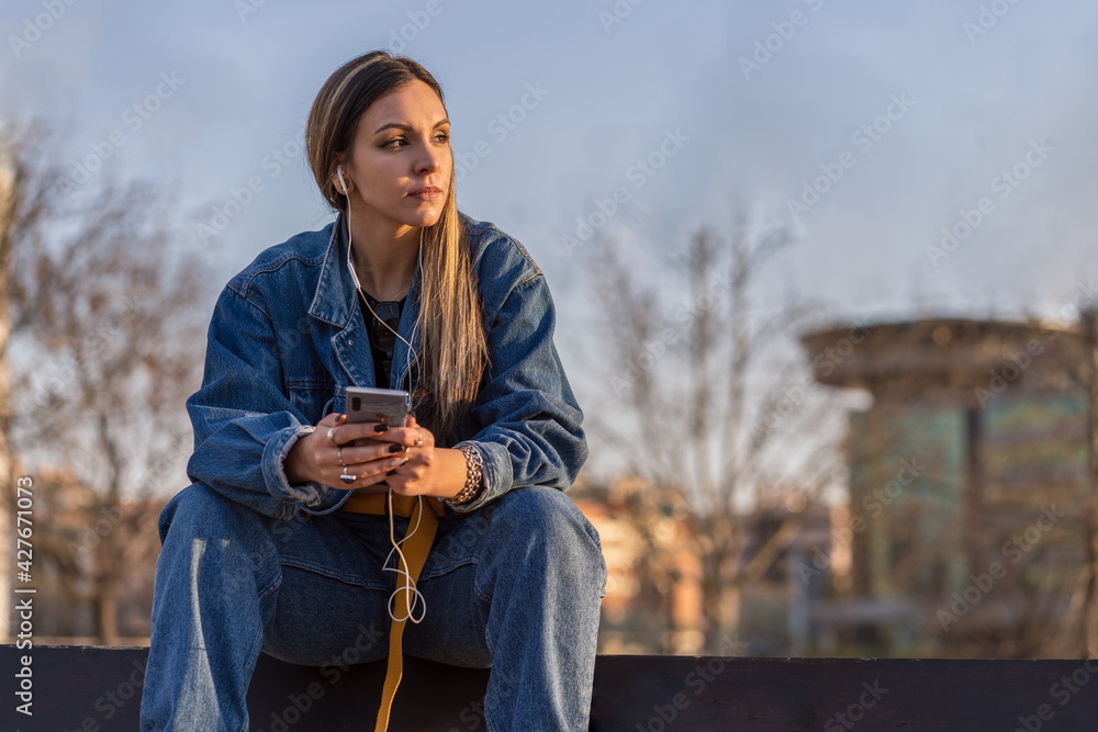 Obraz premium Nice young woman sitting on a bench in a public park with her smartphone