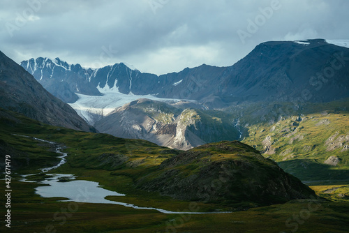 Atmospheric alpine landscape with mountain lake in green valley and glacier under cloudy sky. Awesome highland scenery with beautiful glacial lake among sunlit hills and rocks against mountain range.