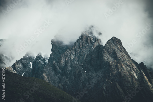 Dark atmospheric surreal landscape with dark rocky mountain top in low clouds in gray cloudy sky. Gray low cloud on high pinnacle. High black rock with snow in low clouds. Surrealist gloomy mountains.