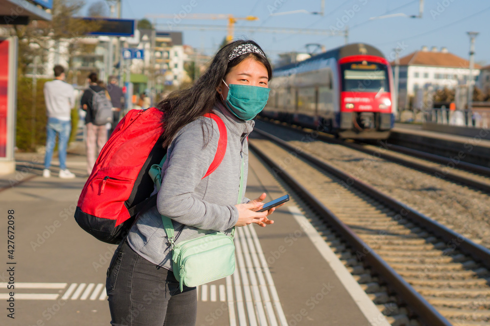 Asian backpacker girl traveling in times of covid19 - young happy and beautiful Korean woman in face mask and backpack waiting for train at station platform visiting Europe