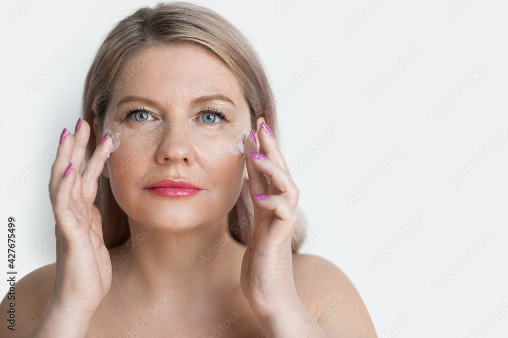 Senior blonde woman is massaging her face with a cream during spa procedures on a white studio wall Senior blonde woman is massaging her face with a cream during spa procedures on a white studio wall
