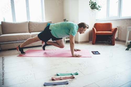 Blonde man is using a tablet while doing fitness exercises at home on a yoga mat © Strelciuc