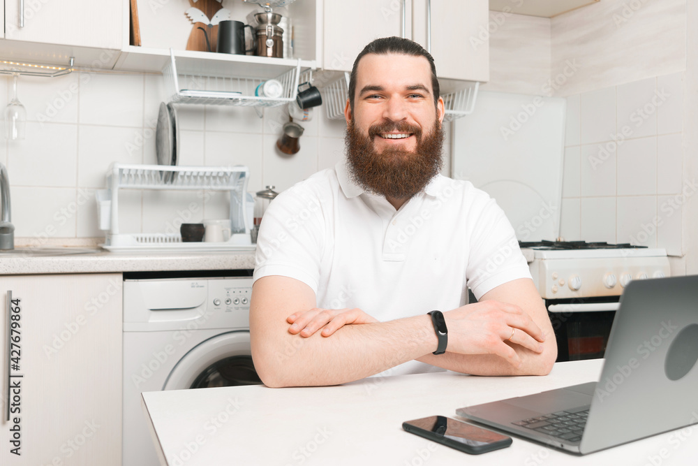 A nice picture of a young bearded man sitting cheerful at a computer at home studying