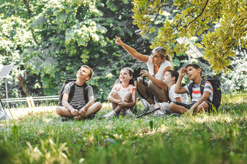 Group of school kids sitting on grass in forest with they teacher.They learning about nature and wildlife.	
