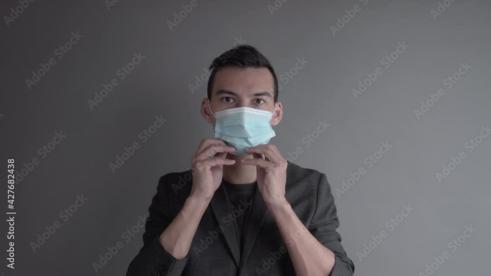 Asian European Guy in a Blazer Puts on a Blue Medical Face Mask Grey Background Medium Shot Asian European Guy in a Blazer Puts on a Blue Medical Face Mask Grey Background Medium Shot