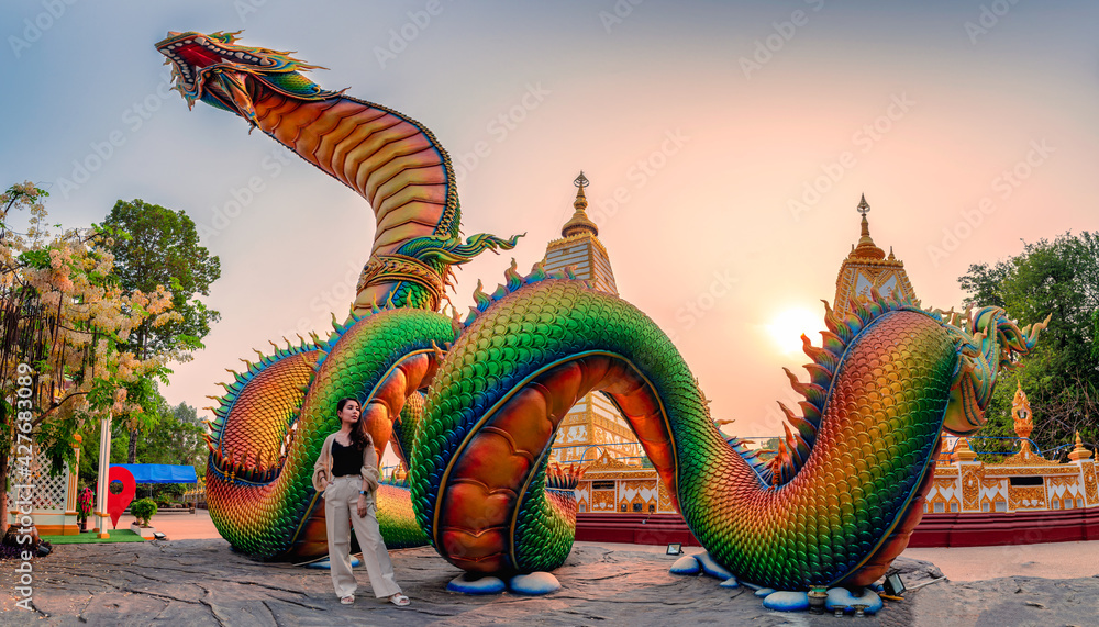 Fototapeta premium Rainbow carve serpent or colorful Thai Naga and asian woman standing in the sunset at Wat Phra That Nong Bua temple