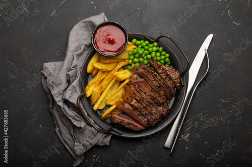 Sliced grilled meat beef steak with potato chips french fries and green peas served with red tomato sauce on rustic metal plate on black stone background table from above 
