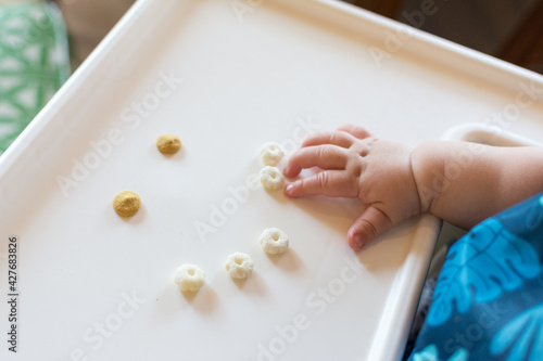 Smiley face made of cereal puffs and yogurt drops on a high chair tray  baby reaching out to self feed