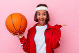 Young mixed race woman playing basketball isolated on pink background showing a copy space on a palm and holding another hand on waist.