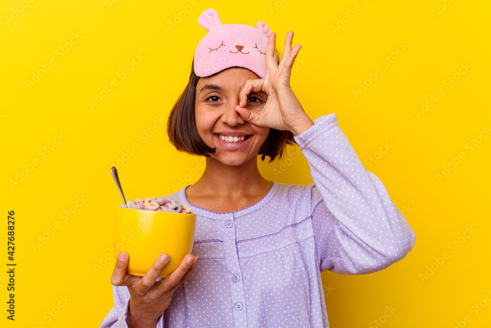 Young mixed race woman eating cereals wearing a pijama isolated on yellow background excited keeping ok gesture on eye. Young mixed race woman eating cereals wearing a pijama isolated on yellow background excited keeping ok gesture on eye.