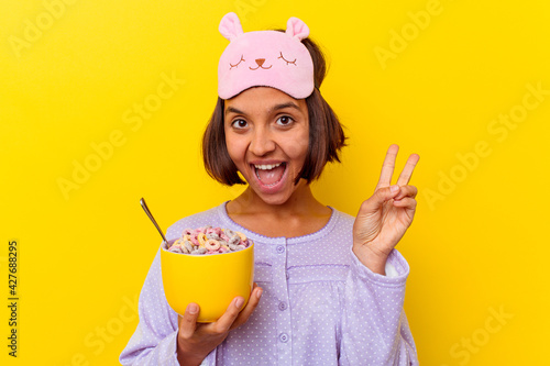Quadro su tela Young mixed race woman eating cereals wearing a pijama isolated on yellow background joyful and carefree showing a peace symbol with fingers