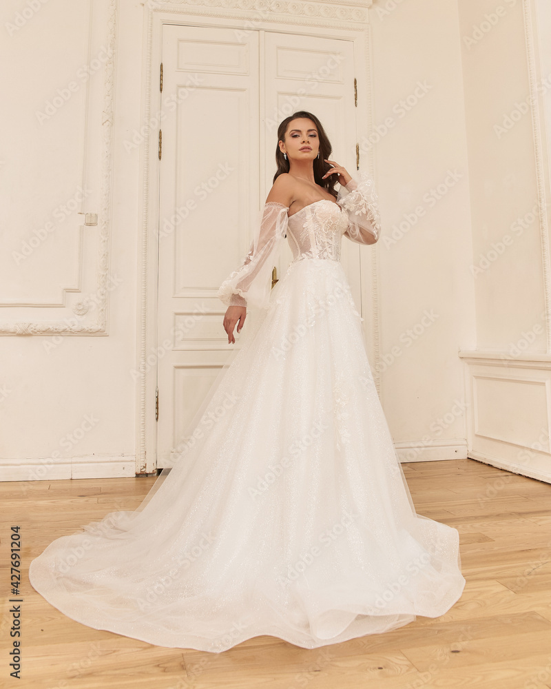 Elegant caucasian woman with natural make-up and hairstyle wearing white bridel dress, standing and posing in bright stylish interior. Full length portrait Elegant caucasian woman with natural make-up and hairstyle wearing white bridel dress, standing and posing in bright stylish interior. Full length portrait