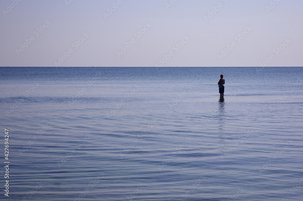 Fisherman fishes in a completely calm sea with his feet covered by water. Fishing in relaxing glass calm sea water Copy space. High quality photo
