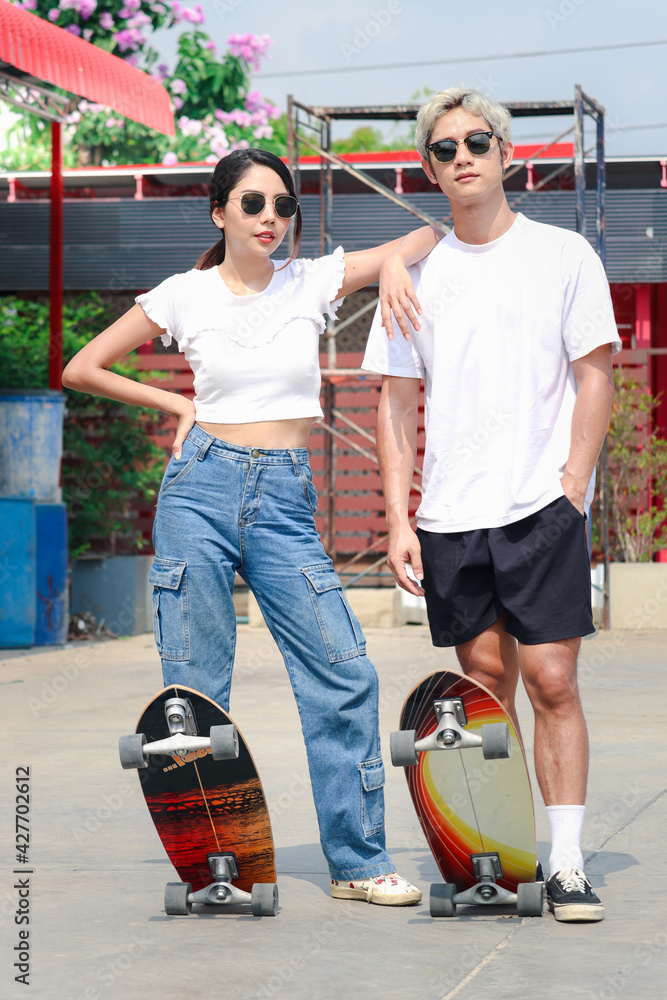 Young Asian man and woman wearing sunglasses with surf skate board on the road outside on sunny day two people enjoy having fun with skateboard extreme sport activity outdoor. Photos Adobe