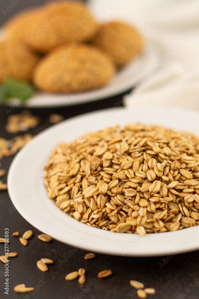 In the foreground, a blurred image of oatmeal in a white plate, in the background oatmeal cookies on a dark background. In the foreground, a blurred image of oatmeal in a white plate, in the background oatmeal cookies on a dark background.