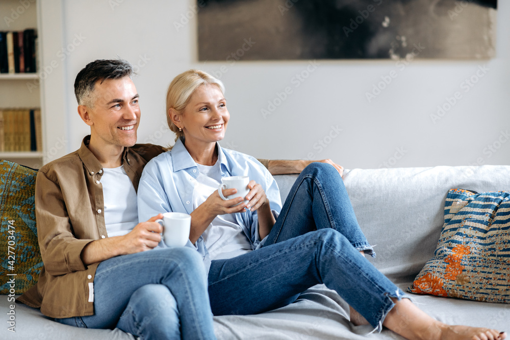 Loving happy adult married caucasian couple in stylish casual clothes, happy husband and wife sitting on the comfortable sofa in the living room, holding cups of tea, chatting, spending time at home