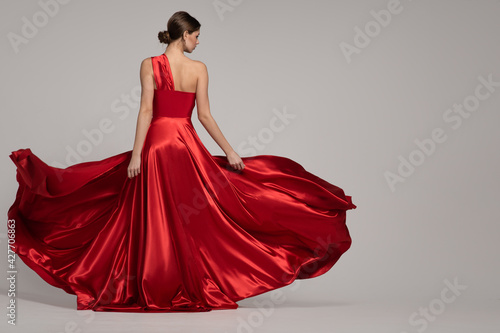 Fototapeta Fashion woman in red long dress on gray background. Back view