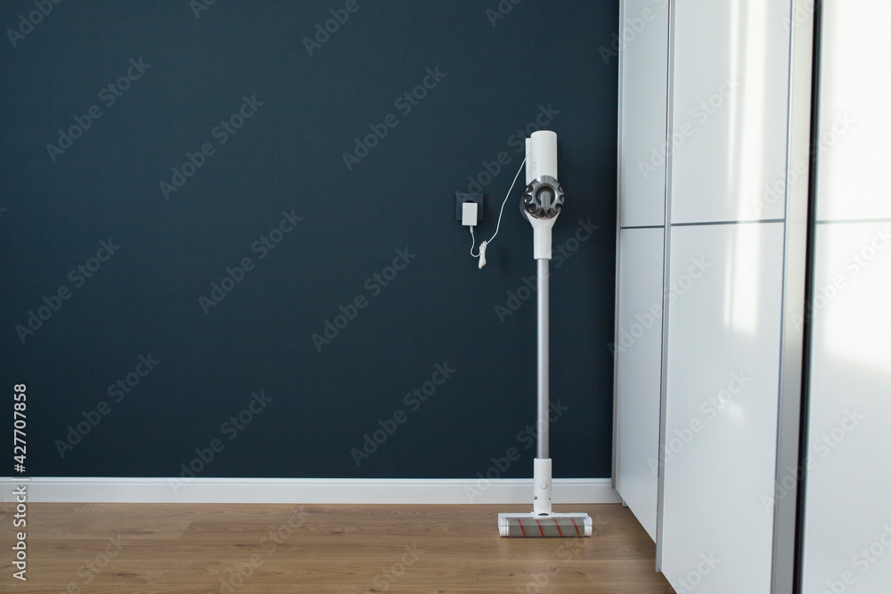 Modern cordless vacuum cleaner charges against a blue wall. Minimalistic interior. - obrazy, fototapety, plakaty 