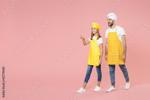 Full length body of teen fun girl dad man father's helper chef cook confectioner baker in yellow apron toque cap uniform walk go point finger aside isolated on pastel pink background studio portrait