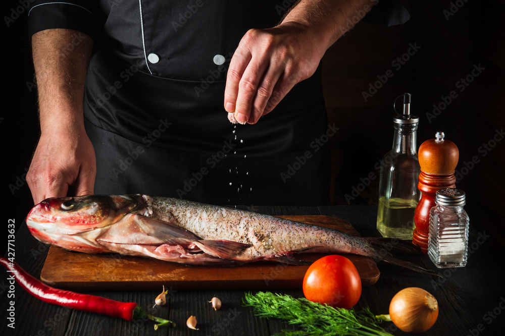 The chef prepares fresh fish bighead carp sprinkling salt with ingredients. Preparing to cook fish food. Working environment in the restaurant kitchen