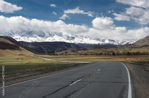 Mountain steppe valley road path with spring green grass and ranges of  snow mountains rocks on a horizon skyline under blue sky © nighttman