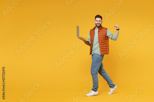Young freelancer happy caucasian man in orange vest mint sweatshirt glasses using laptop pc computer chat online browsing internet do winner gesture clench fist isolated on yellow background studio.