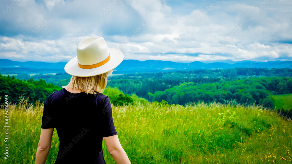 Blonde woman in black dress and white hat at meadow with mountains on background