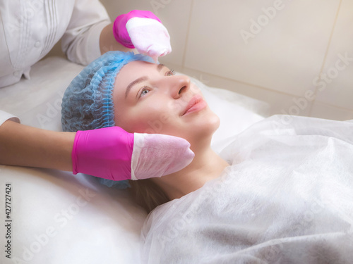 Professional cosmetologist is cleansing skin with pads. Hands in pink gloves of beautician and caucasian woman close up. Cleaning stage. Facial skin care. Beauty salon, cosmetology clinic, spa.