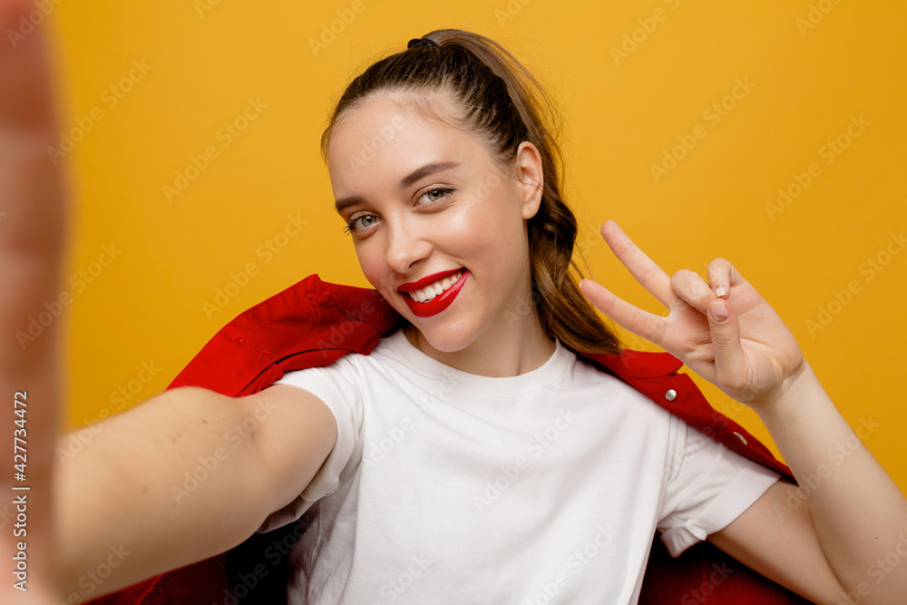 Smiling happy girl with red lips wearing red jacket making selfie with peace sign over isolated backdrop Smiling happy girl with red lips wearing red jacket making selfie with peace sign over isolated backdrop