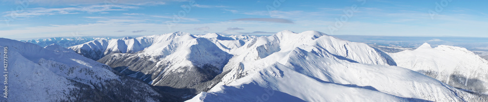 Breathtaking panoramic shot of mountain peaks covered in snow during the winter season. A blue sky with few white clouds can be seen in the background. Zakopane - Kasprowy Wierch