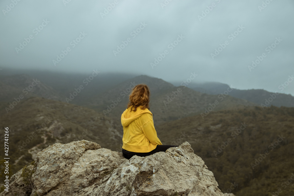 Woman from behind with a yellow jacket sits on a rock on top of a mountain in autumn with mist