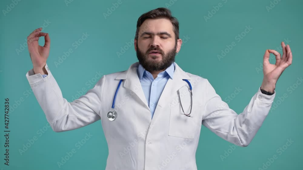 Calm doctor in white medical coat relaxing, meditating. Man calms down, breathes deeply with mudra om on blue studio background. Calm doctor in white medical coat relaxing, meditating. Man calms down, breathes deeply with mudra om on blue studio background.