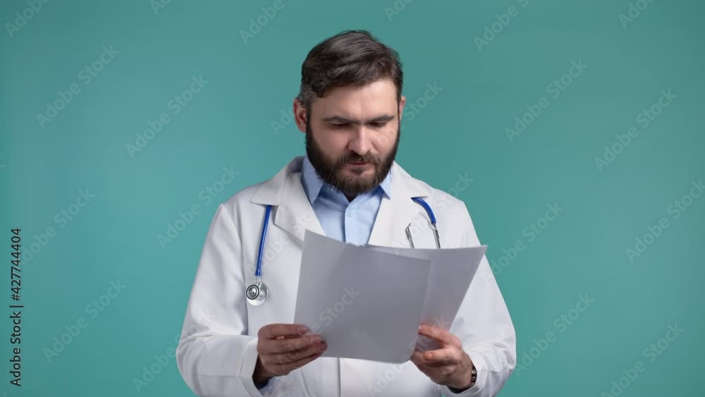 Satisfied man in professional medical coat holding files papers isolated on blue background. He nods his head approvingly to diagnosis Doctor with beard and stethoscope. Satisfied man in professional medical coat holding files papers isolated on blue background. He nods his head approvingly to diagnosis Doctor with beard and stethoscope.