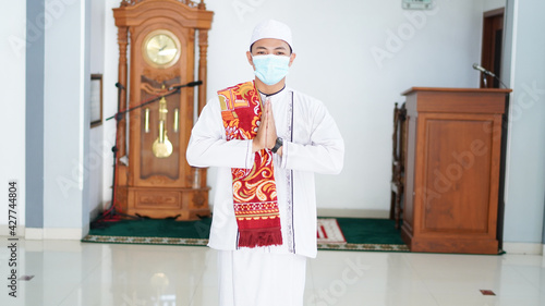 A portrait of an asian muslim man stands up in a greetting pose to Namaste hands, welcoming guests, Ied Fitr greeting at mosque. Wearing mask