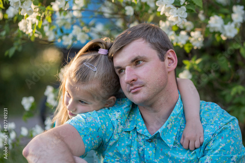 Father and daughter in the flowering white trees. A girl with blue eyes hugs the shoulders of a blond dad in a blue shirt. They sit before the branches of a flowering Jasmine.
