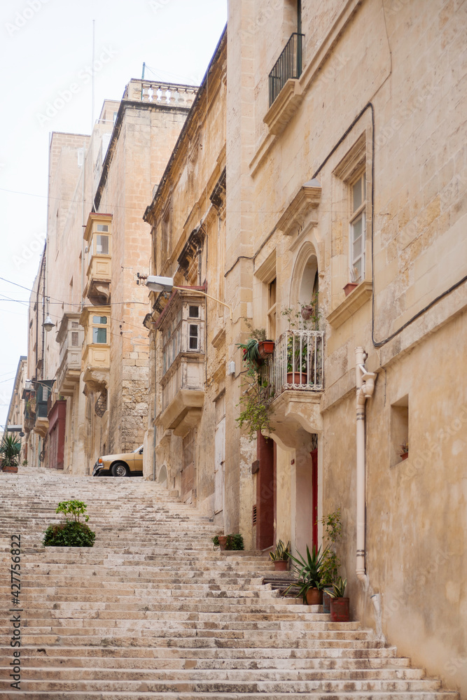 Ancient narrow street in Valletta, Malta. Limestone buildings with old fashioned lanterns and balconies. Staircase with many steps as pavement.