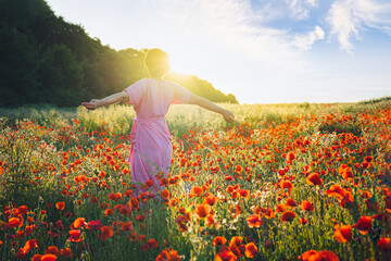 Backview happy young woman in a pink dress with raised arms relaxing in red poppies flowers meadow in sunset light. A simple pleasure for mental health. Nature relaxation. Selective focus. Copy space