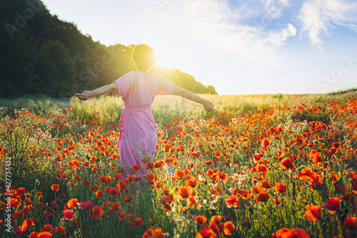 Backview happy young woman in a pink dress with raised arms relaxing in red poppies flowers meadow in sunset light. A simple pleasure for mental health. Nature relaxation. Selective focus. Copy space