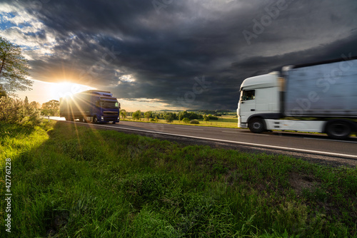 Obraz na płótnie Motion blurred white truck and blue truck driving on the asphalt road in rural landscape in the rays of the sunset with dark storm cloud w salonie