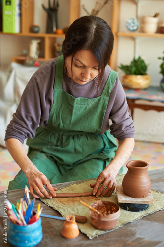 Woman master of pottery in a green apron sculpts a jug of red clay