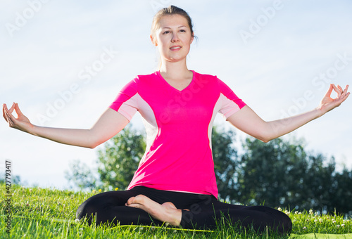 Portrait of sportswoman which is sitting and meditating in pink T-shirt in the park.