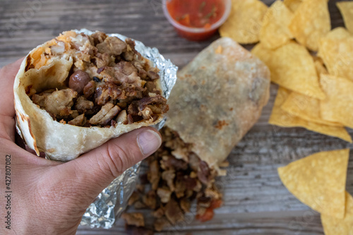 Hand holding a carne asada beef burrito wrapped in foil over a table with chips and salsa at a Mexican food restaurant.