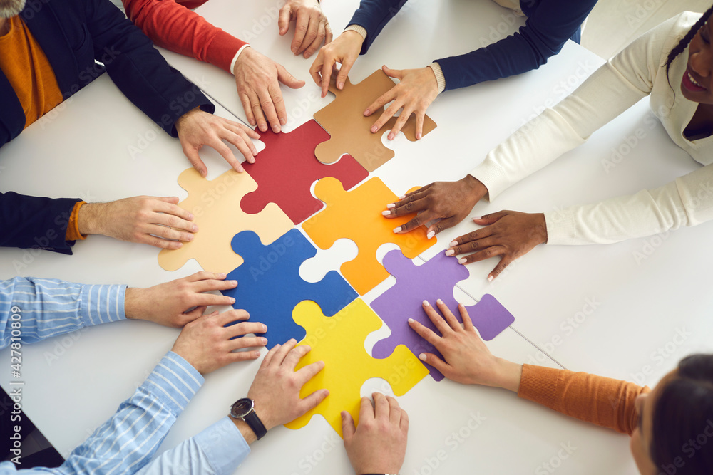 © Studio Romantic - Solidarity, strategy collaboration, business workflow and communication research concept. Diverse multiethnic office people team assembling jigsaw puzzle pieces holding hands on table top view
