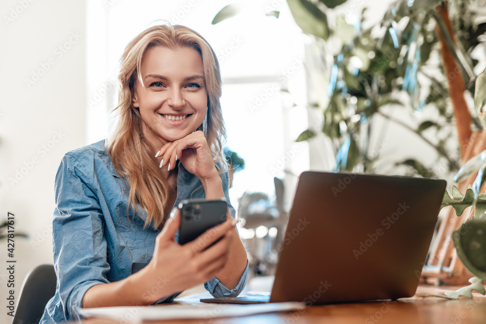 Glad blond haired woman sitting at table with laptop and mobilephone Glad blond haired woman sitting at table with laptop and mobilephone