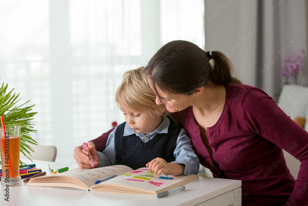 Cute preschool child, blond boy, filling some homework in a work book and coloring, mother helping him Cute preschool child, blond boy, filling some homework in a work book and coloring, mother helping him