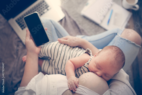 Cute caucasian mom and newborn baby, mom breastfeeds her baby and works with phone and laptop. Top view.