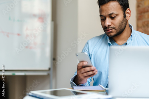 Middle eastern unshaven man using mobile phone while working with laptop