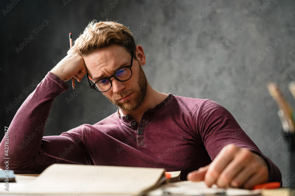 The portrait of tired man resting his head with his hand while sitting at the table in the studio The portrait of tired man resting his head with his hand while sitting at the table in the studio