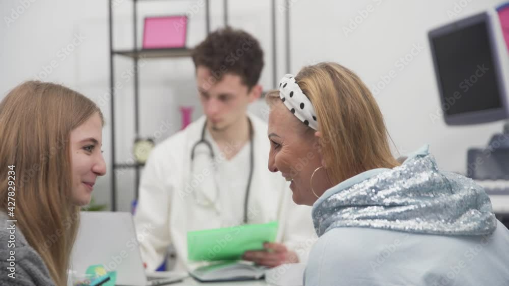The daughter and mother are happily chatting in the office while the doctor prepares the medical records The daughter and mother are happily chatting in the office while the doctor prepares the medical records